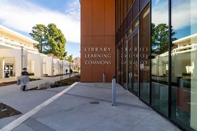 Exterior view of a modern library building with glass windows and a sign that reads 'Library Learning Commons' in an area with trees and a clear blue sky.