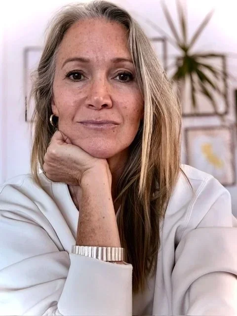 A woman with long blonde hair, wearing a white top and gold hoop earrings, resting her chin on her hand, in a room with art on the wall and a potted plant in the background.