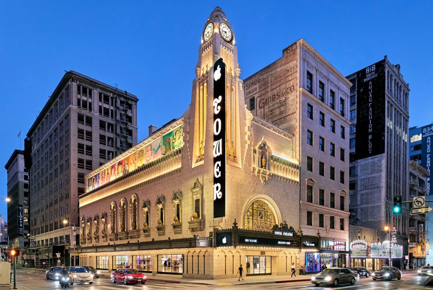 The Tower Theater in Los Angeles illuminated at dusk, with cars driving by and other buildings in the background.