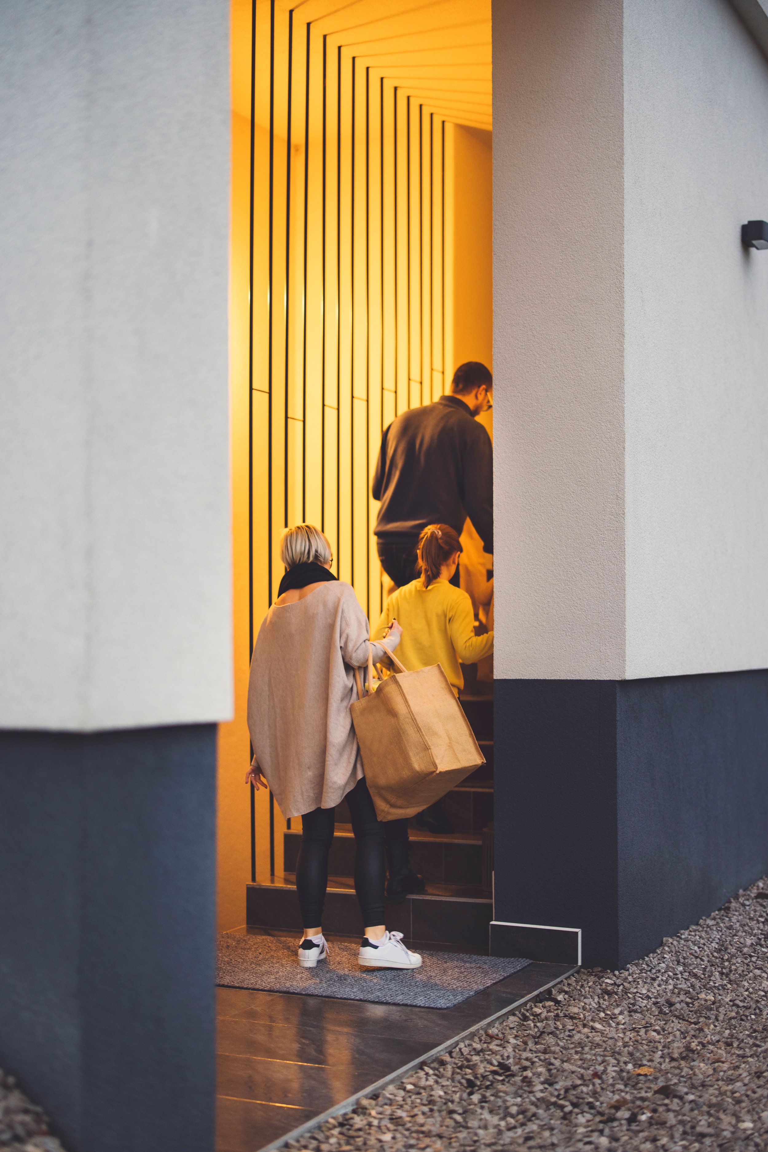 People entering a building through a doorway, seen from outside. The building has a dark exterior with a textured white wall and a decorative yellow and black interior wall visible through the doorway.