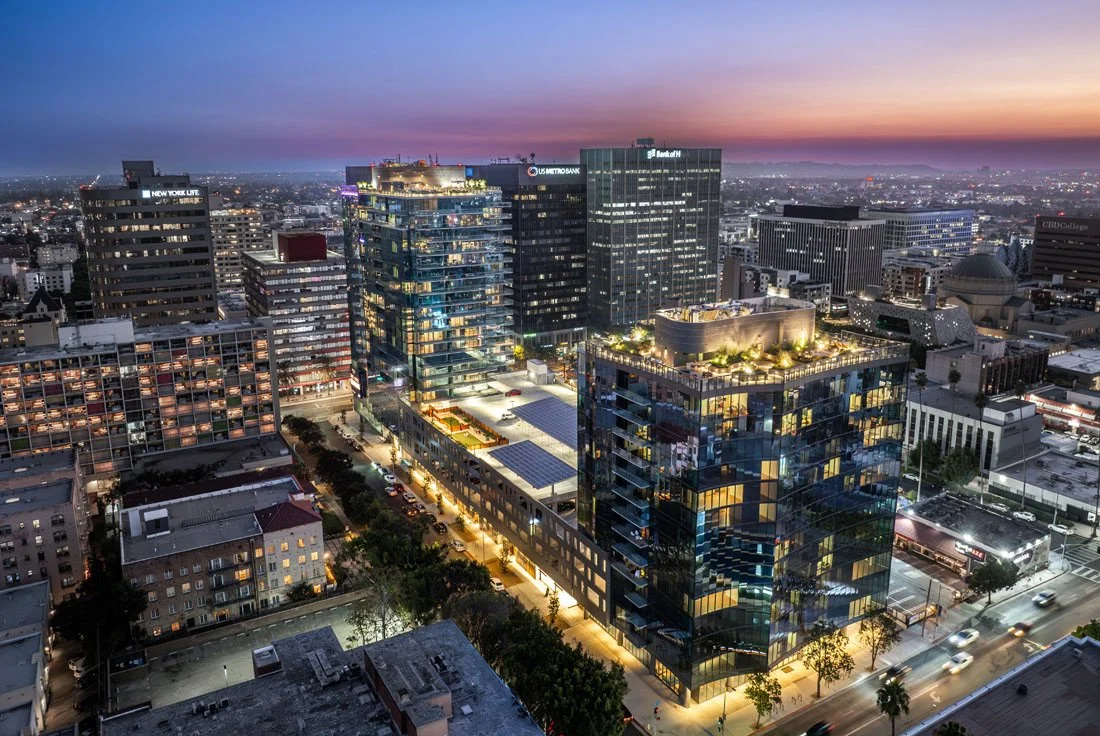 A high-angle view of a city skyline at dusk with illuminated modern glass office buildings and a sunset sky.