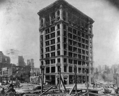 A black and white photo of a partially destroyed multi-story building in an urban area, with debris and damaged surrounding structures.