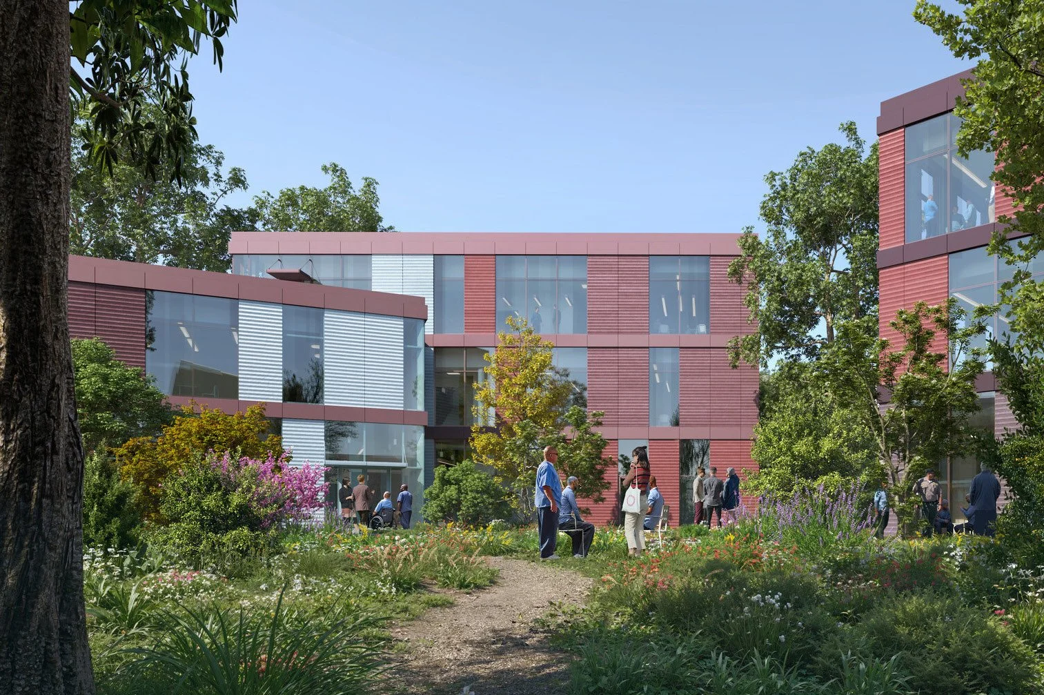 People walking and sitting in a green courtyard with trees, colorful flowers, and a modern red brick building with large glass windows in the background.