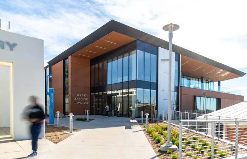 Modern library building with large glass windows and a brown exterior, labeled 'Library Learning Commons', with students entering and walking outside on a sunny day.