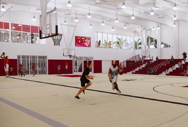 People playing basketball on an indoor court with red and white walls, a scoreboard, and seating areas.