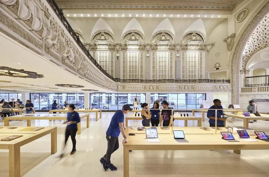 Inside an Apple Store with displays of iPads and customers browsing, featuring ornate architecture with high ceilings and decorative columns.