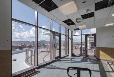 Empty interior space with large glass windows showing a cityscape and mountains in the background, sunlight streaming in.