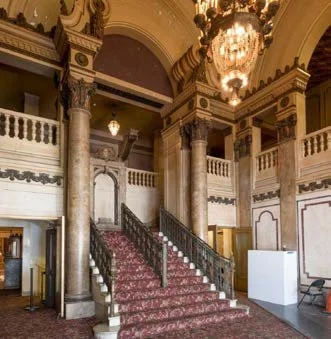 Elegant staircase with red carpet in grand historic building interior with ornate columns and chandelier.