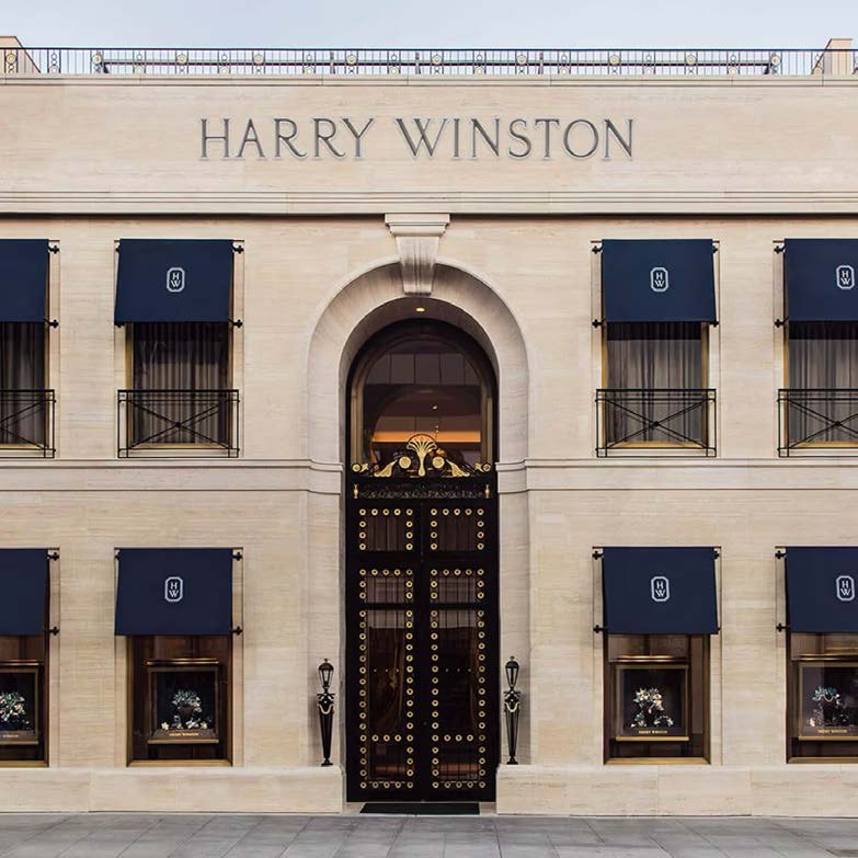 Facade of Harry Winston store with black doors, beige stone walls, and blue awnings over windows.