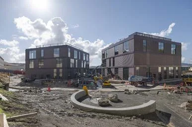 Construction site with two modern black and gray buildings, construction equipment, and materials, under a partly cloudy sky.