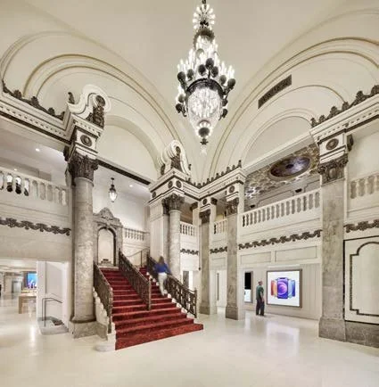 Elegant interior of a grand building with a wide staircase covered in a red carpet, a large chandelier hanging from the ceiling, and ornate architectural details including columns and moldings.