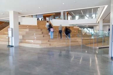 Interior view of a modern building with wide wooden stairs and glass railings, with people walking and looking around.