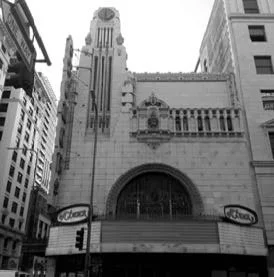 Black and white photo of a tall, ornate building with a clock tower, surrounded by other tall buildings on a city street.
