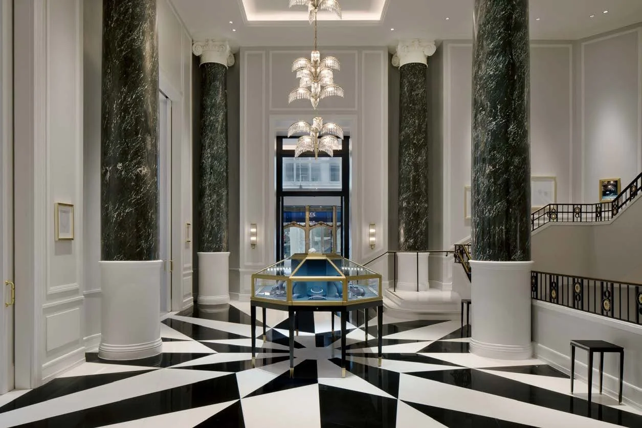 Elegant lobby with black and white geometric patterned floor, tall black marble columns with white capitals, large chandelier, glass display case in center, staircase with black and gold railing to the right, and window at the back.