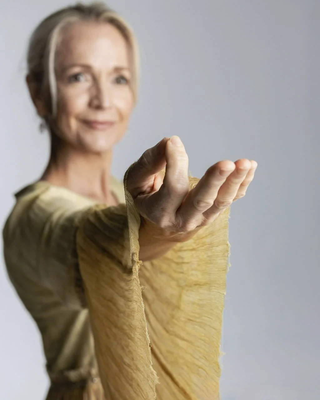 A woman with short gray hair wearing a yellowish top, extending her hand forward, smiling softly, against a plain gray background.