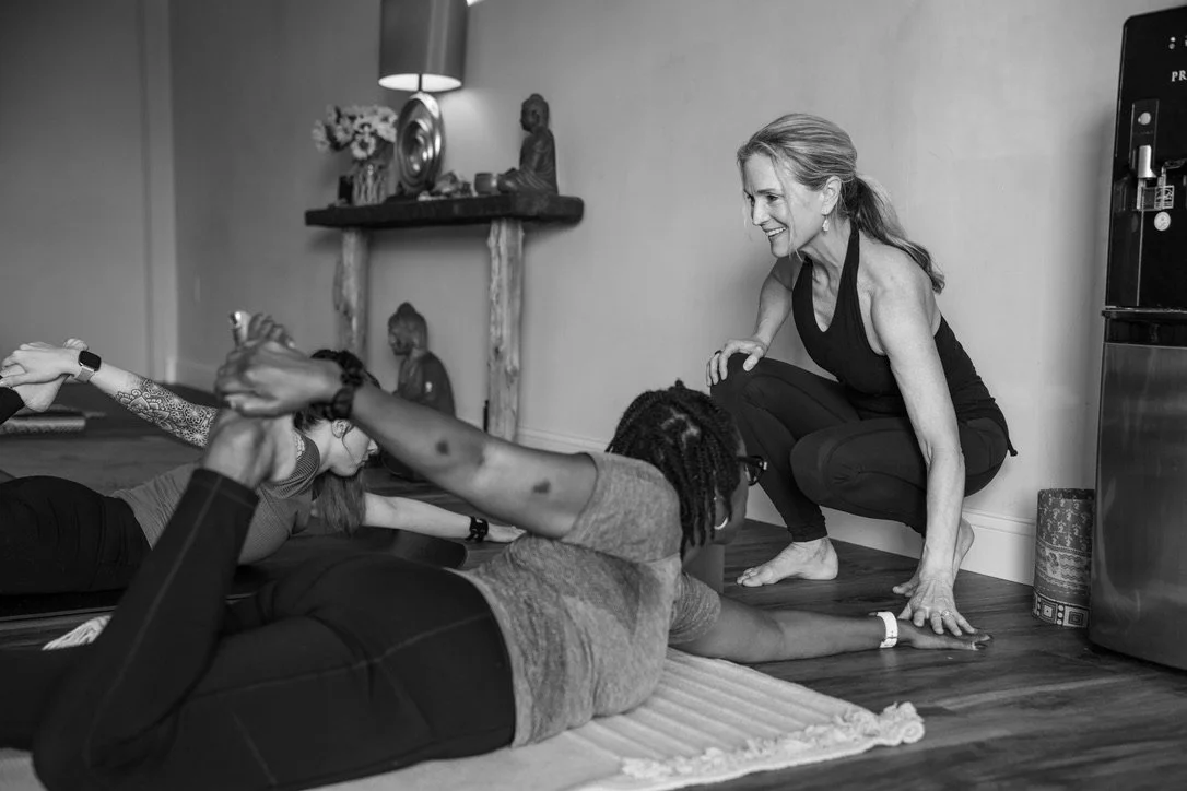 Pamela teaching a yoga class, gently adjusting a student, in a cozy indoor space with a Buddha statue, flowers, and decorative items on a shelf in the background.