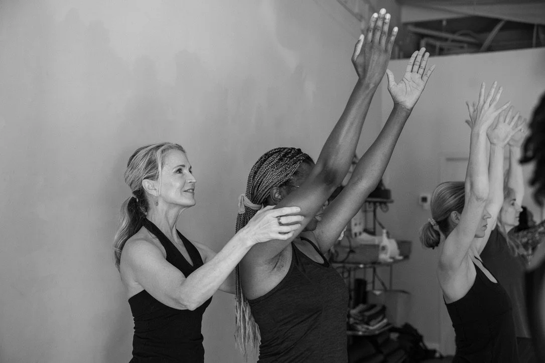 Pamela Meriwether assisting a student in yoga class, while other students practice in the background..