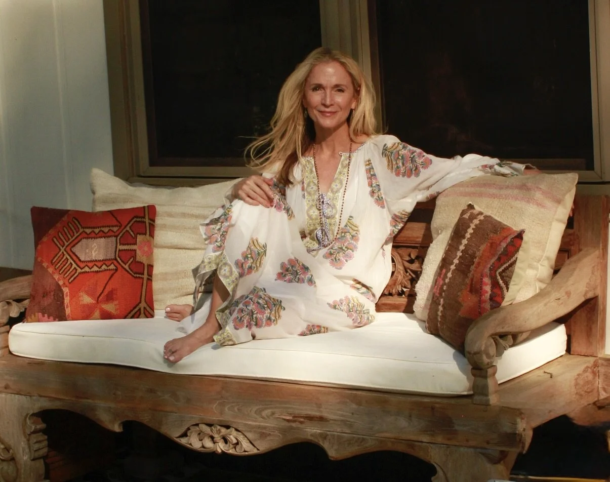 Pamela sitting on a sofa with cream cushions and decorative pillows, wearing a white embroidered dress.
