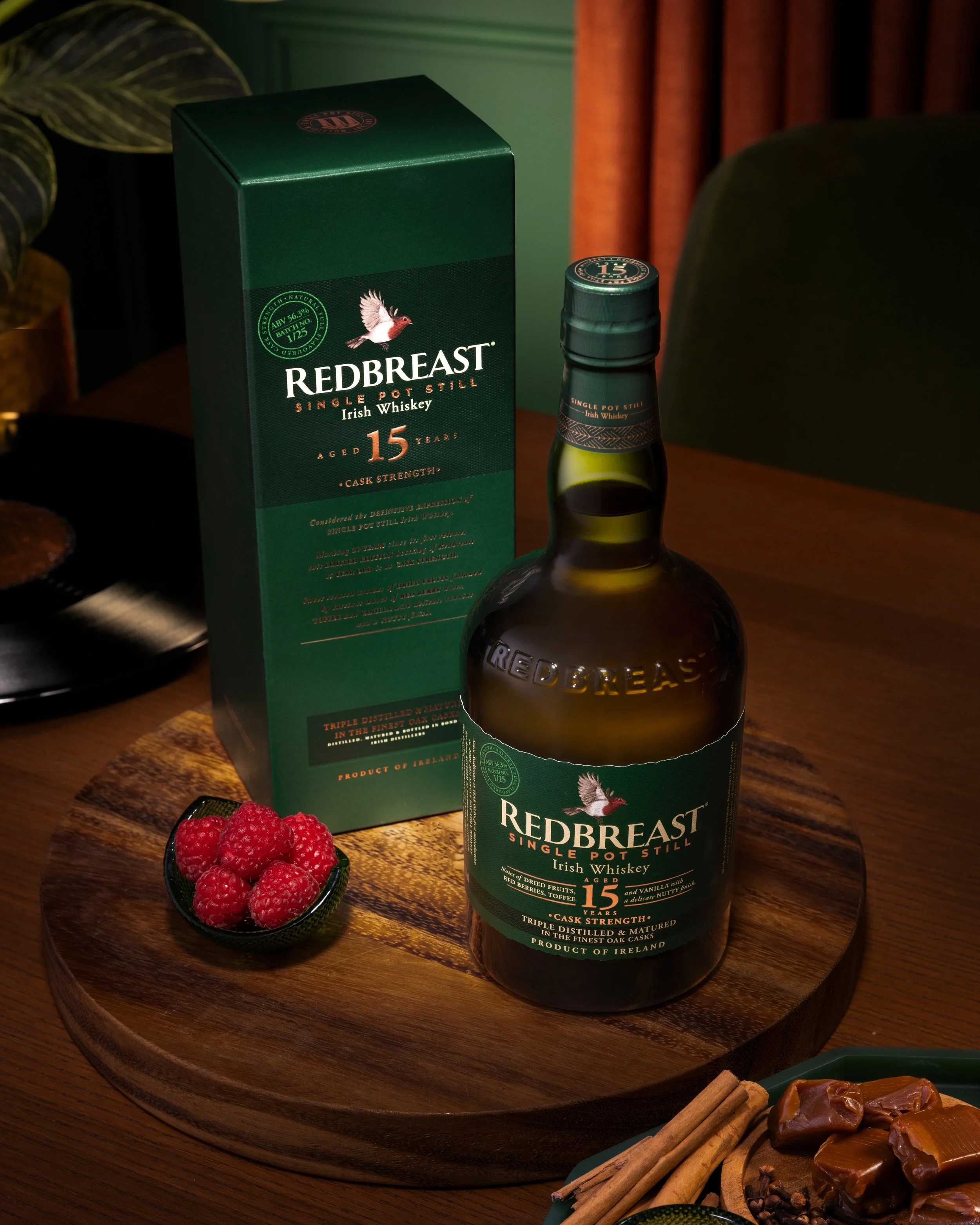 A bottle of Redbreast 15-year-old Irish whiskey with its green packaging box on a wooden tray. A small bowl of raspberries is also on the tray, along with cinnamon sticks and caramel candies in the foreground.