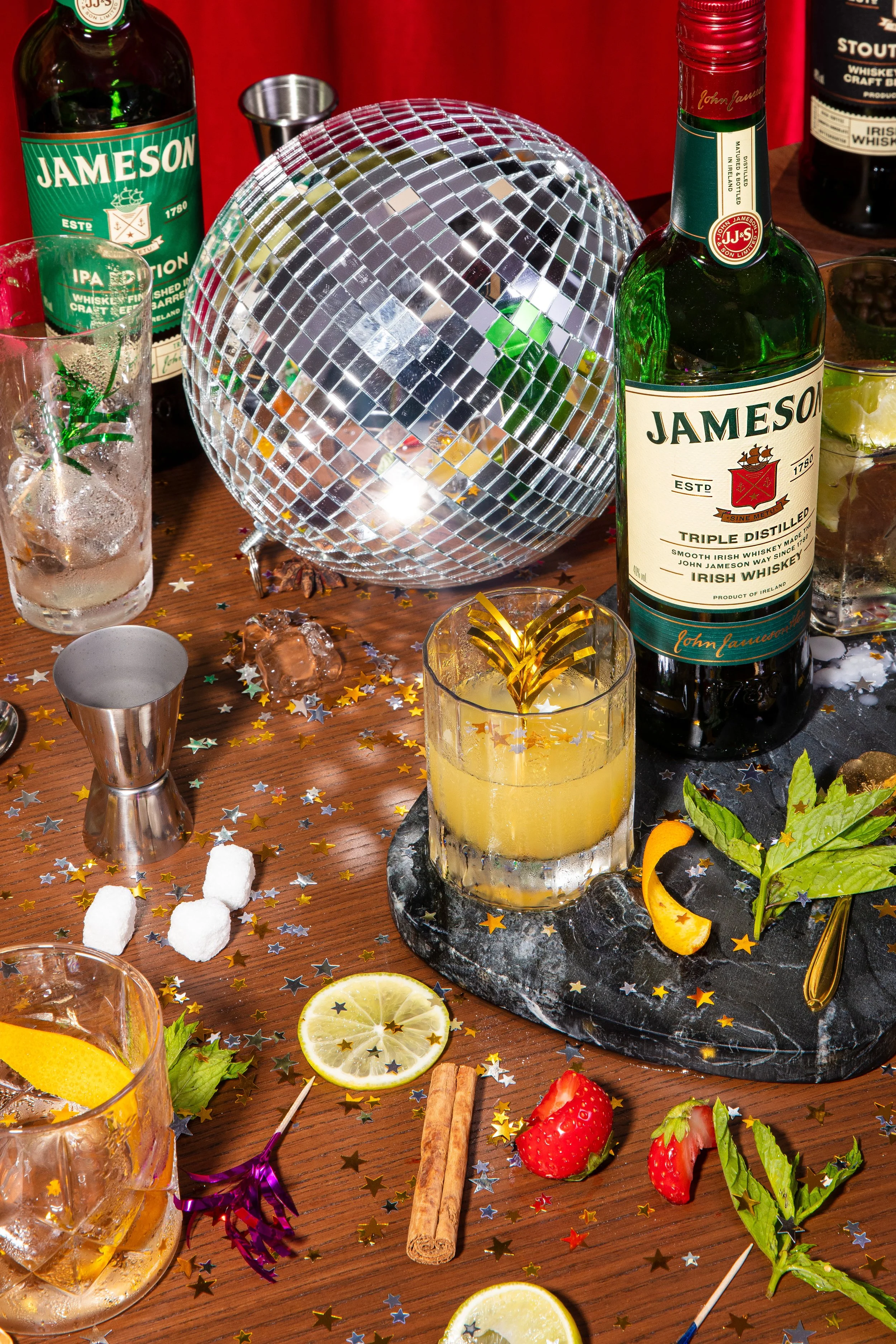 Party scene with drinks, including whiskey bottles, a disco ball, lemon slices, strawberries, mint leaves, and festive decorations on a wooden table.