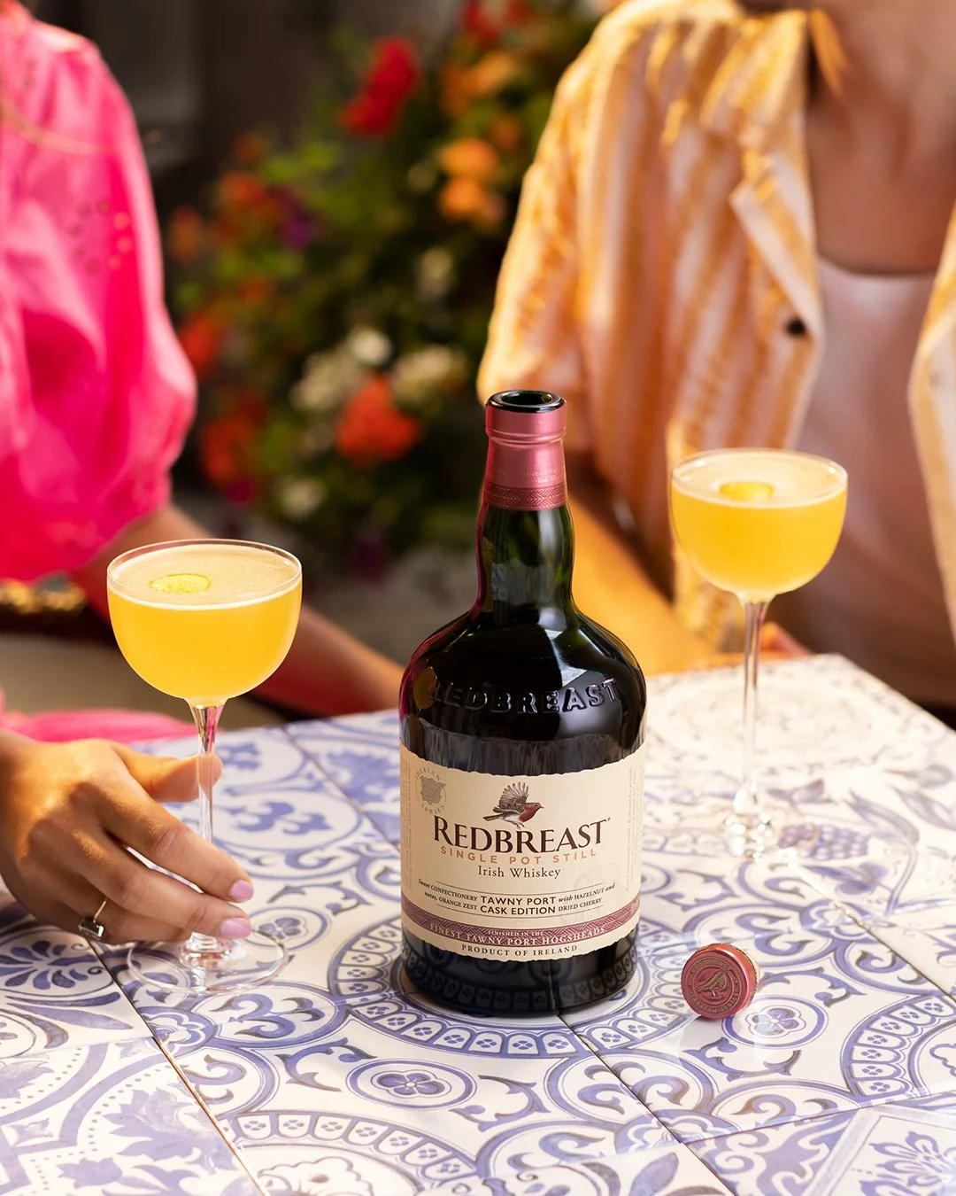 Two women sitting at a table with a blue and white patterned tablecloth, with a bottle of Redbreast Irish whiskey and two yellow cocktails in coupe glasses.
