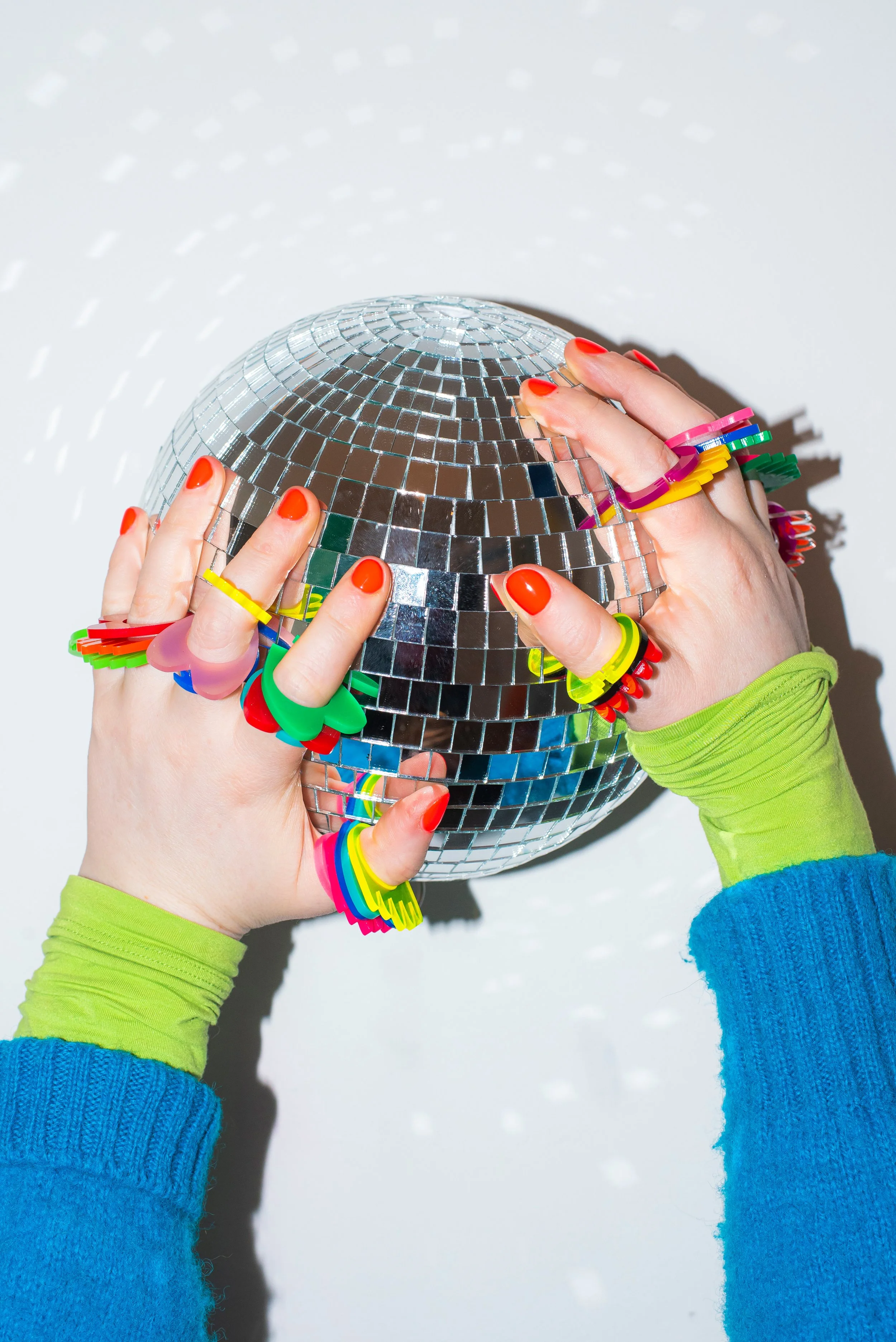 Person wearing colorful jewelry and painted nails holds a small disco ball against a white background, with bright shadows cast.