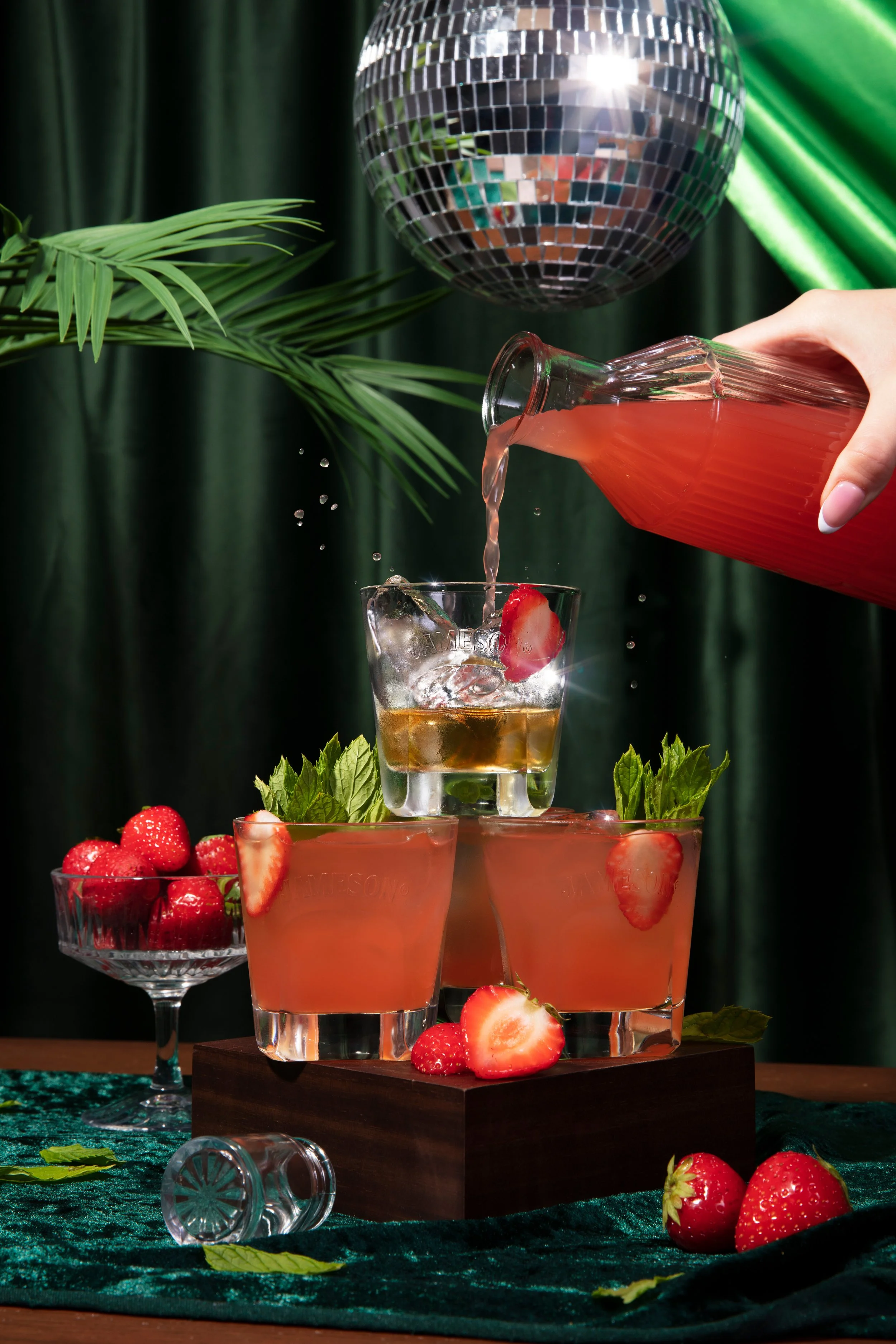 A glass of pink strawberry cocktail being poured, with strawberries and mint leaves around it, a bowl of strawberries, and a disco ball in the background, set on a table with a dark green fabric and a wooden stand.