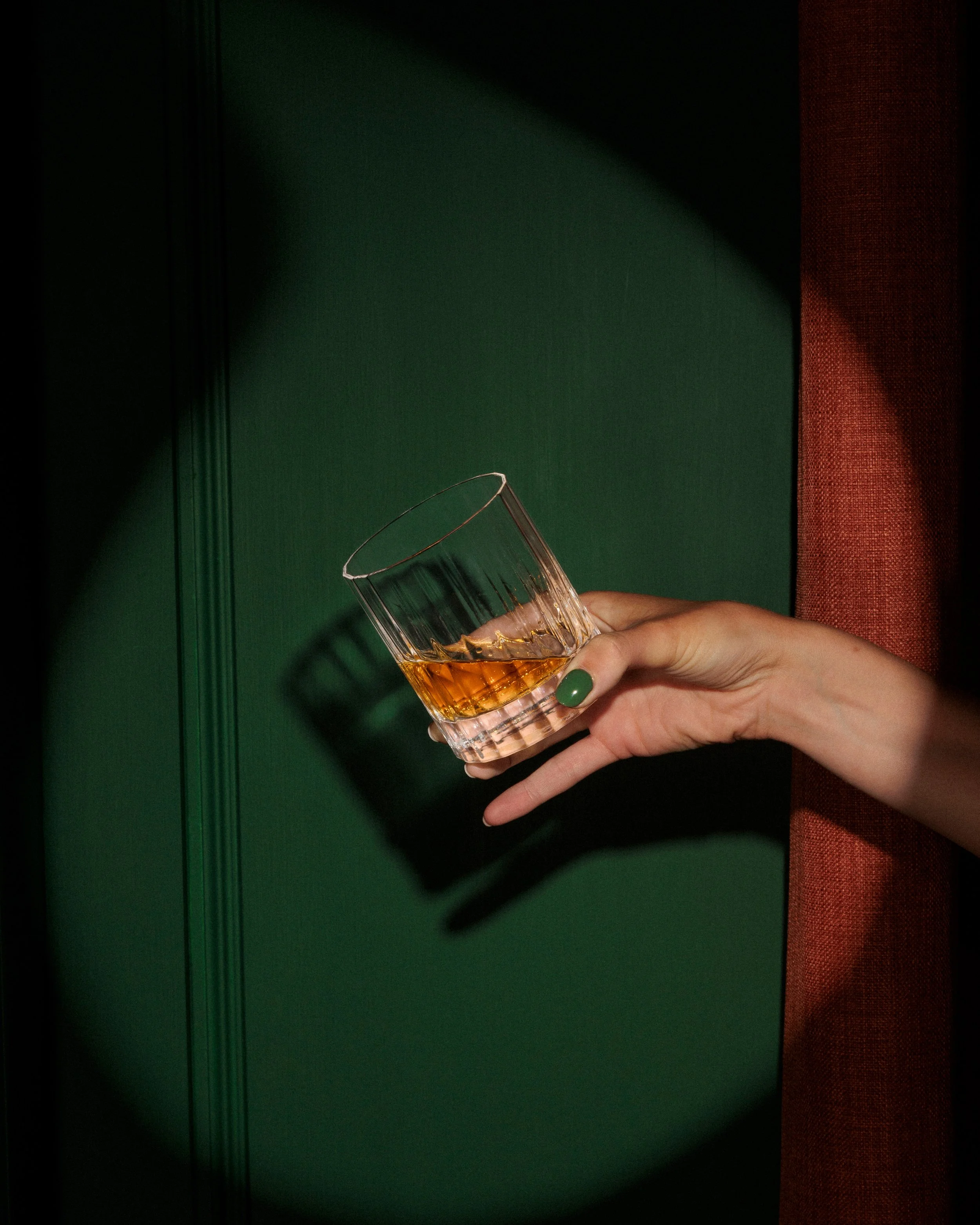 Person holding a glass of amber-colored beverage, with green-painted door and red curtain in the background.