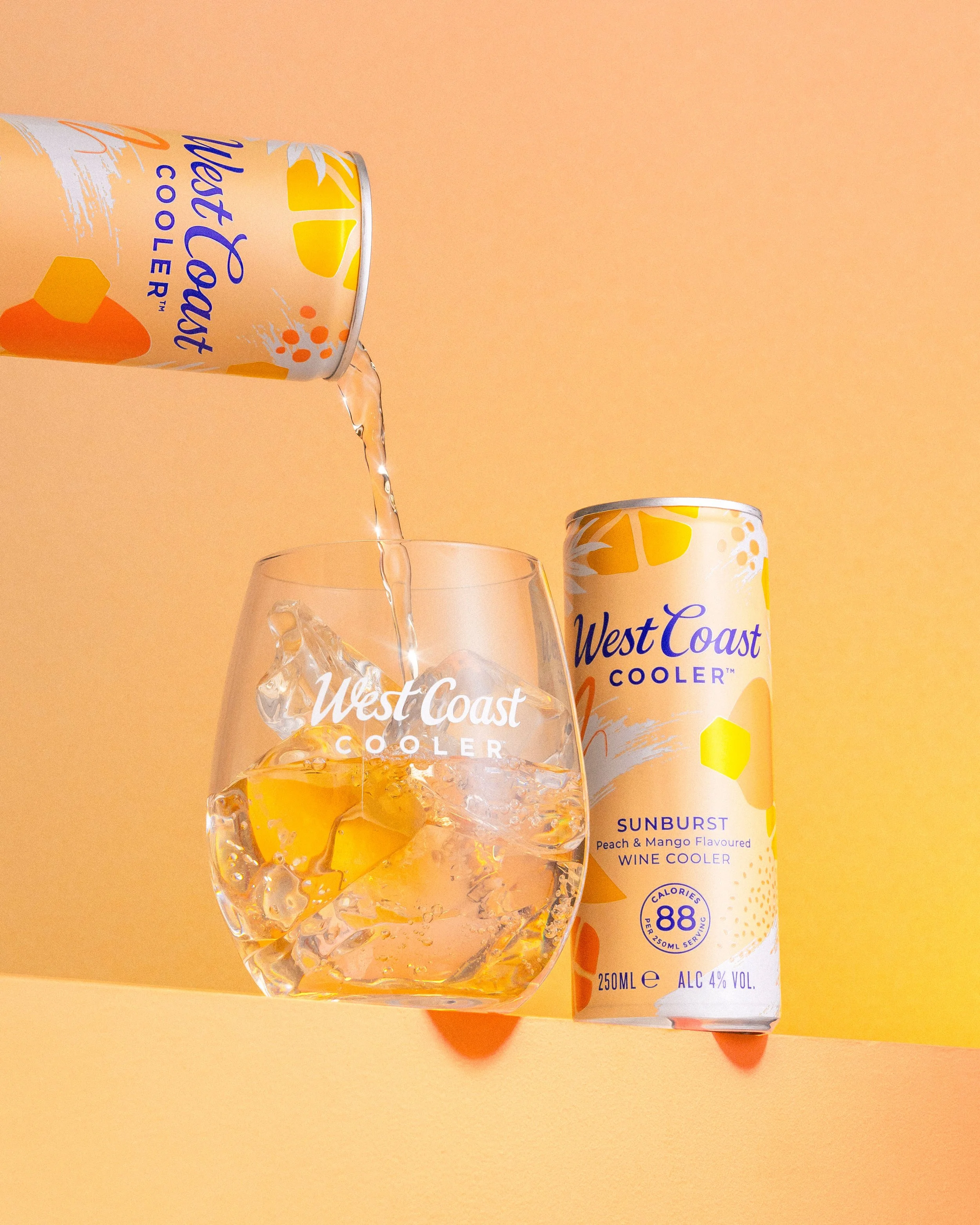 A can of West Coast Cooler Sunburst Peach and Mango flavored wine cooler tilted, pouring liquid into a glass with ice. The can is white with orange, yellow, and purple accents, and the glass is transparent with the West Coast Cooler logo and ice cube