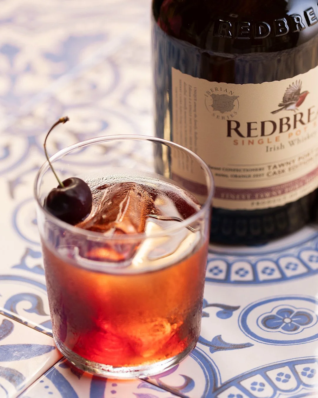 A glass of red cherry-flavored cocktail with ice and a cherry on top, placed on a patterned tablecloth, with a bottle of Redbreast Irish whiskey in the background.