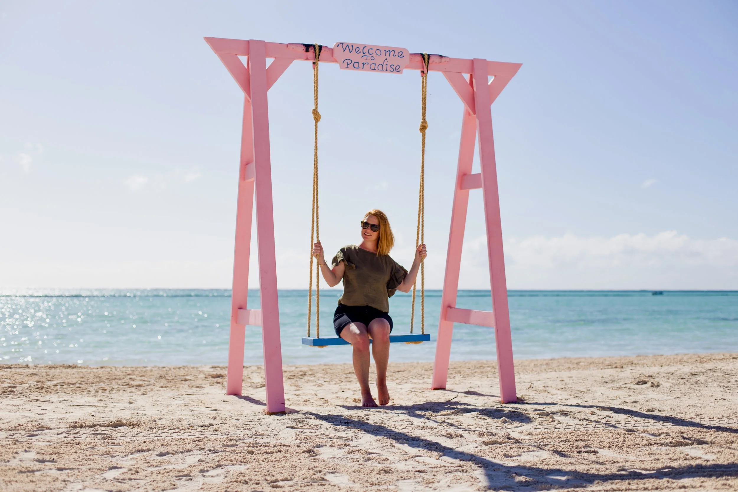 Woman with red hair sitting on a blue swing hanging from a pink wooden frame on the beach, with ocean and sky in the background. The swing has a sign that says "Welcome to Paradise."
