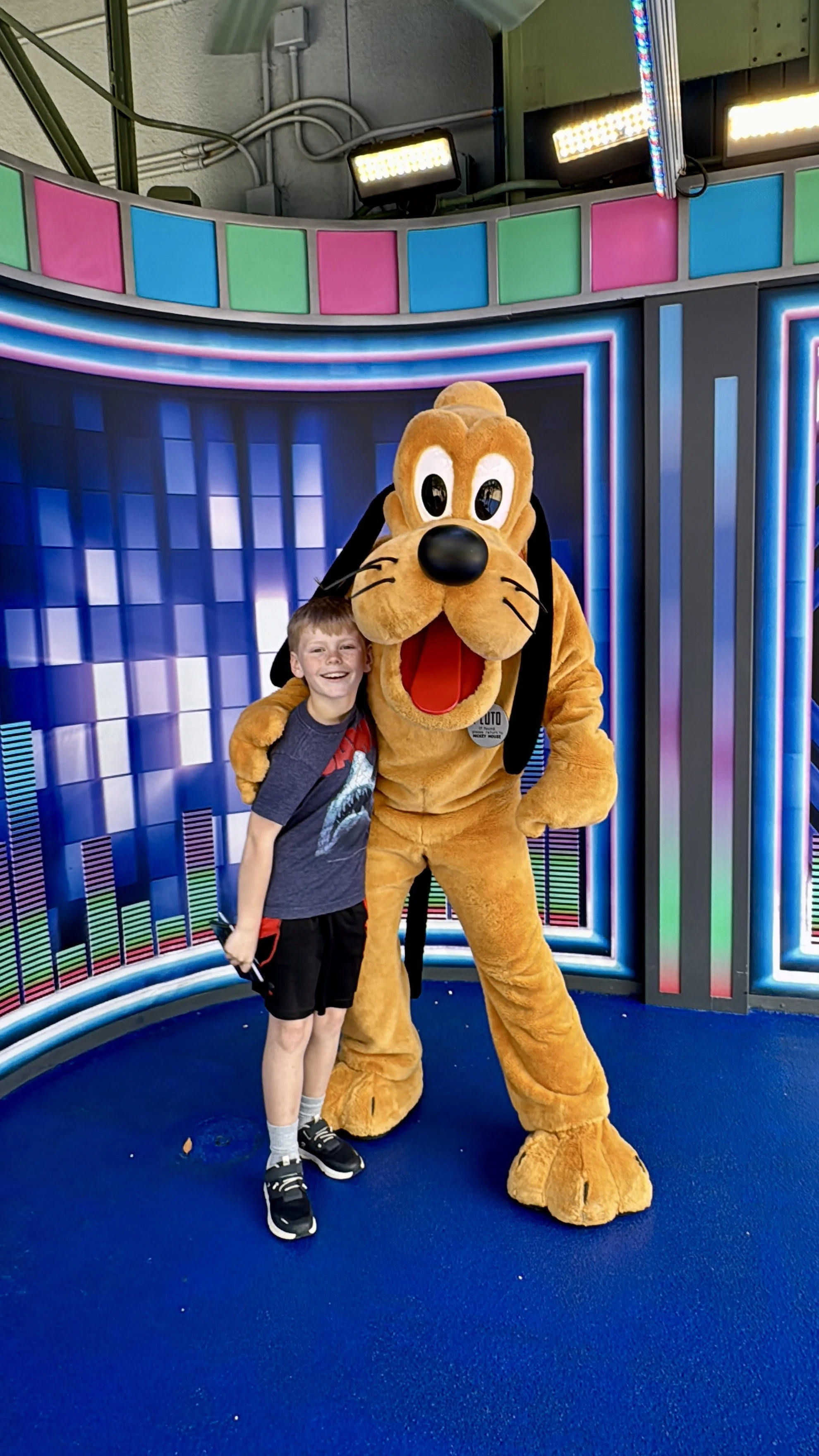 A young boy smiling and hugging Pluto, on a Disney vacation, while posing on a blue carpet with a colorful, animated digital backdrop and stage lights overhead.