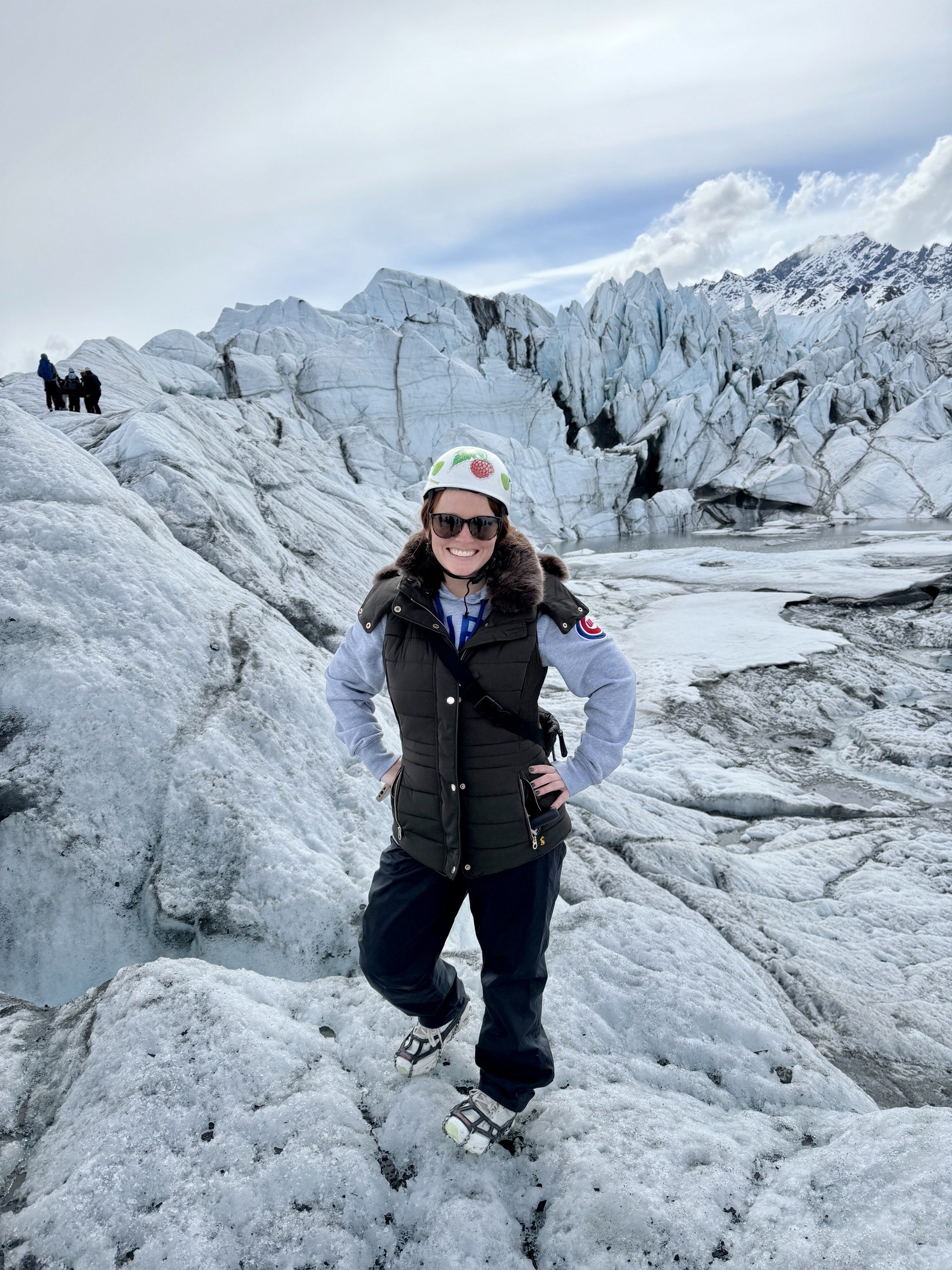 A woman wearing sunglasses, a black vest, and a helmet stands on ice in front of a glacier, smiling at the camera.