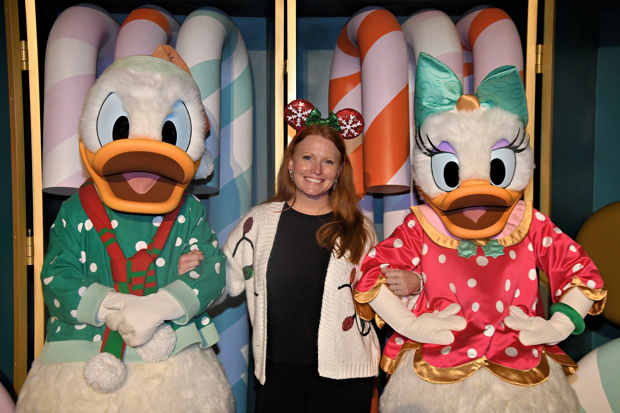 A woman standing between Donald Duck and Daisy Duck dressed in Christmas attire, with colorful candy cane decorations in the background.