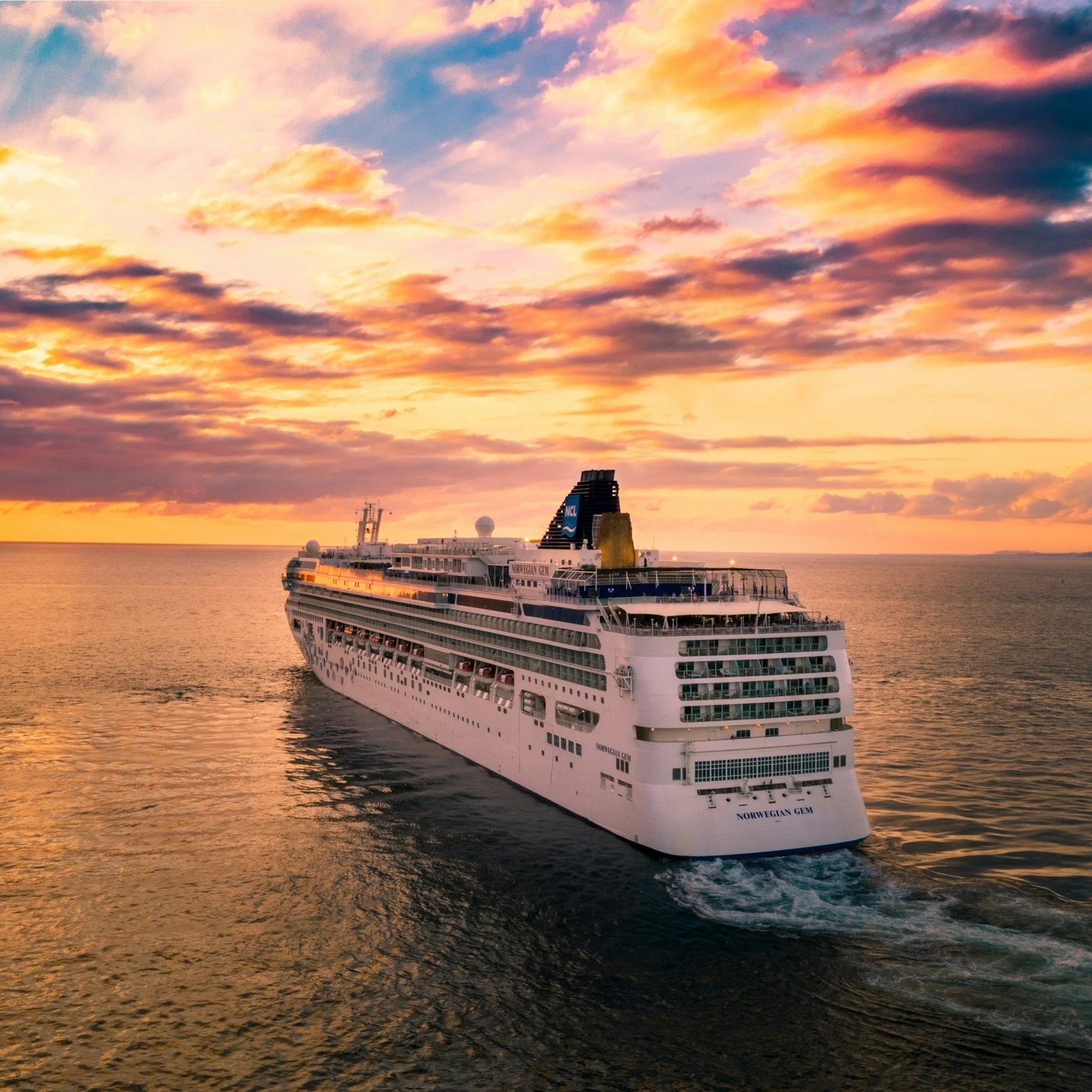 A large cruise ship named Norwegian Gem sailing in the ocean at sunset with a colorful sky overhead.