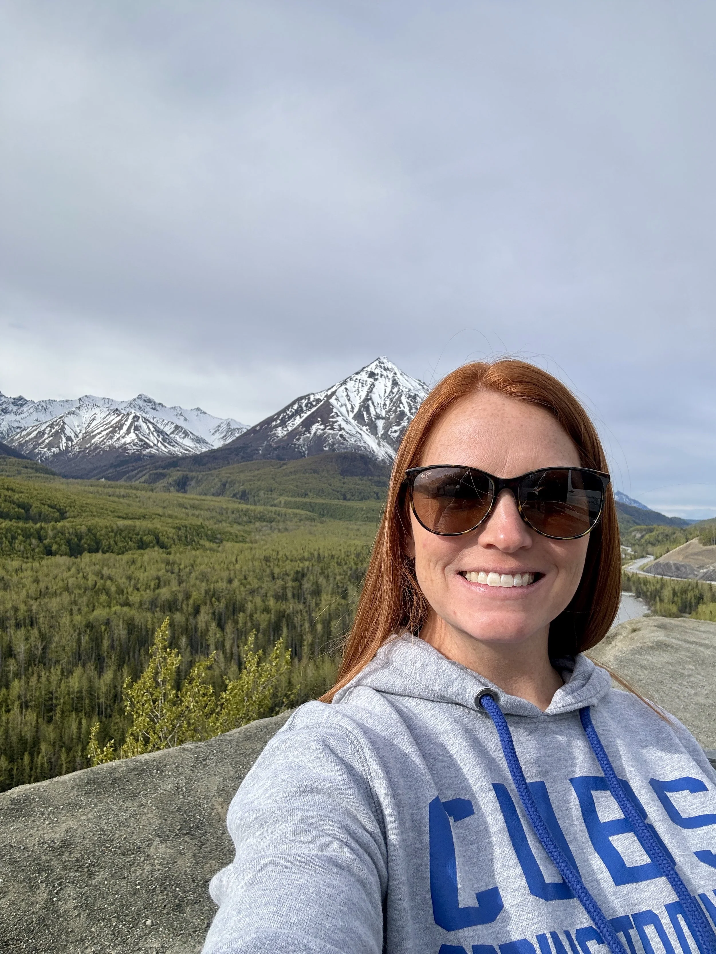 Travel agent, Samantha Harris, taking a selfie with a forested mountain landscape in the background, including snow-capped peaks.
