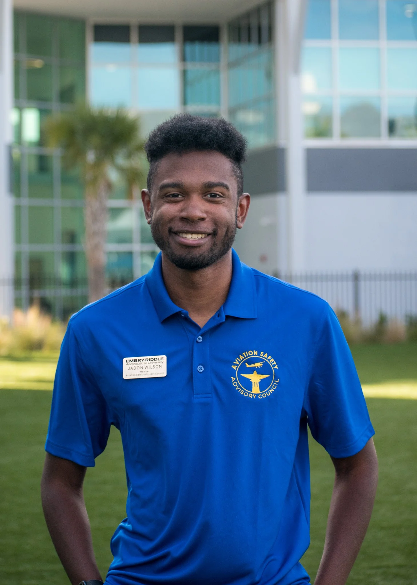 A young man with dark hair and a beard wearing a blue polo shirt with an aviation safety advisory council logo and a name tag, smiling outdoors in front of a modern glass building and greenery.