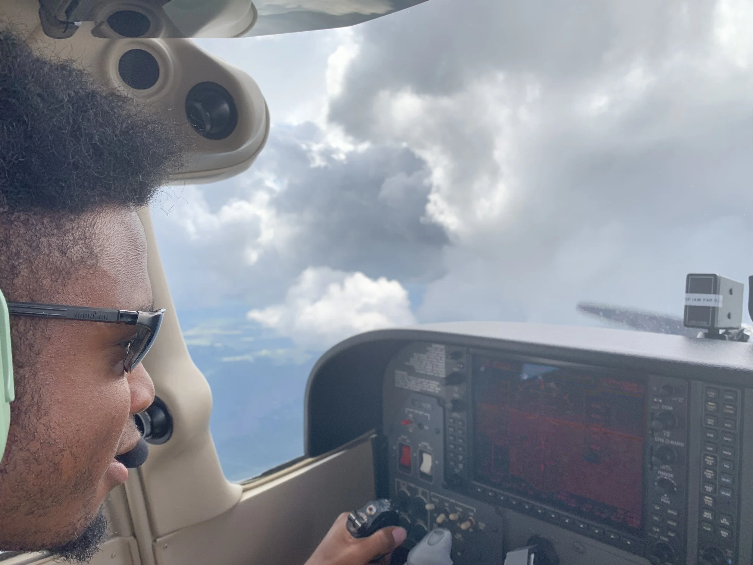 A man with facial hair and glasses is sitting in the cockpit of an airplane, looking down at controls. Outside the window, the sky is cloudy.