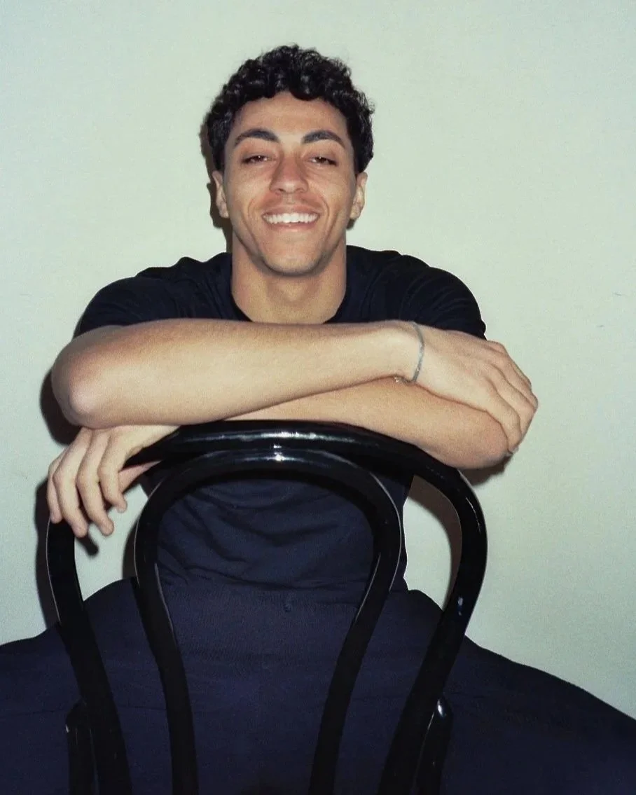 Giovane uomo seduto con braccia appoggiate a una sedia nera, sorridente davanti a una parete bianca