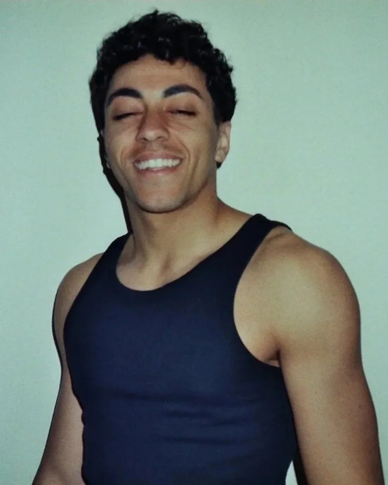 Uomo con capelli ricci, sopracciglia folte, sorriso ampio, vestito con canottiera nera, sfondo neutro.