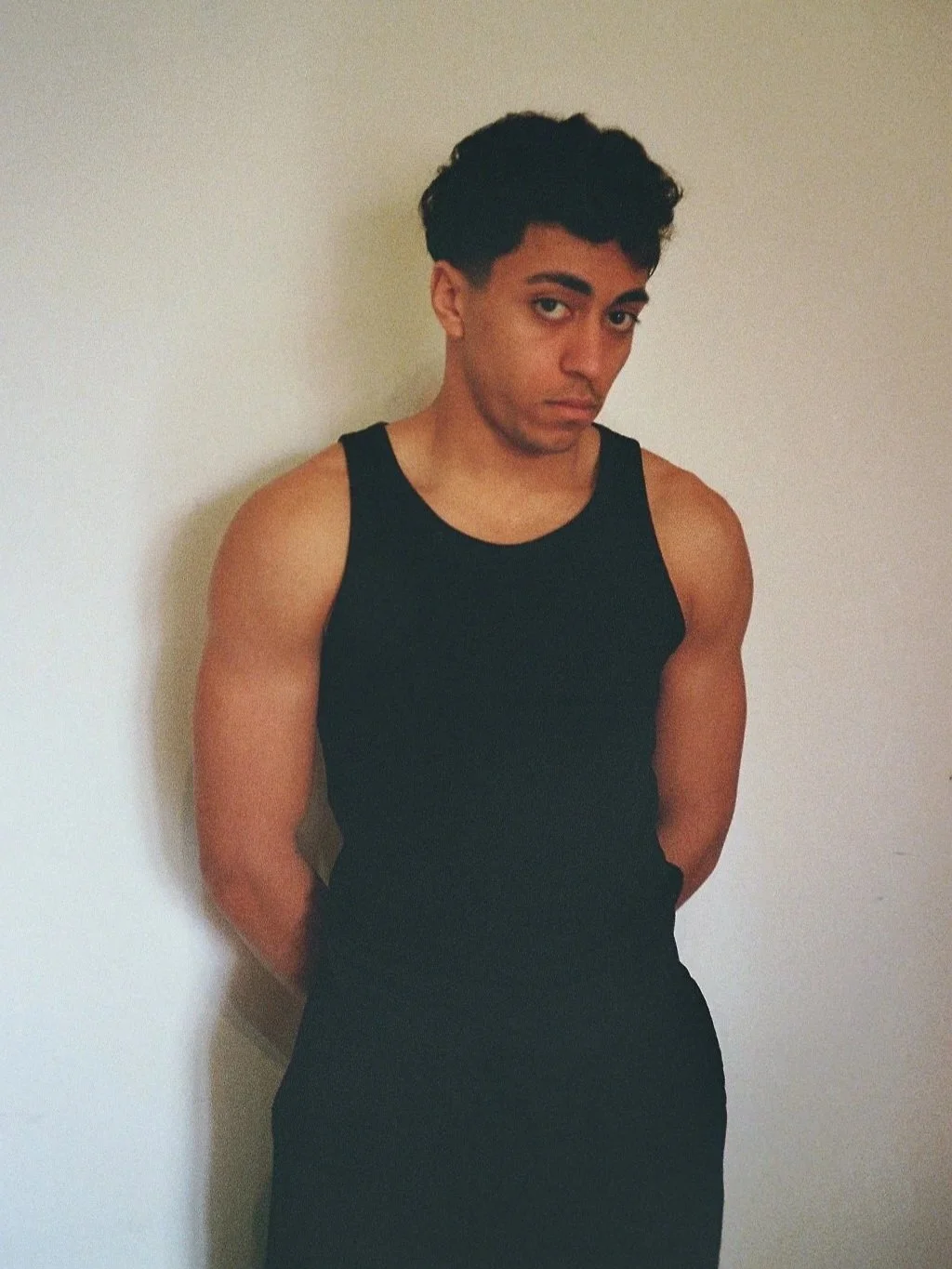 Un giovane uomo con i capelli ricci scuri indossa una canotta nera e sta contro un muro bianco, con le mani dietro la schiena.