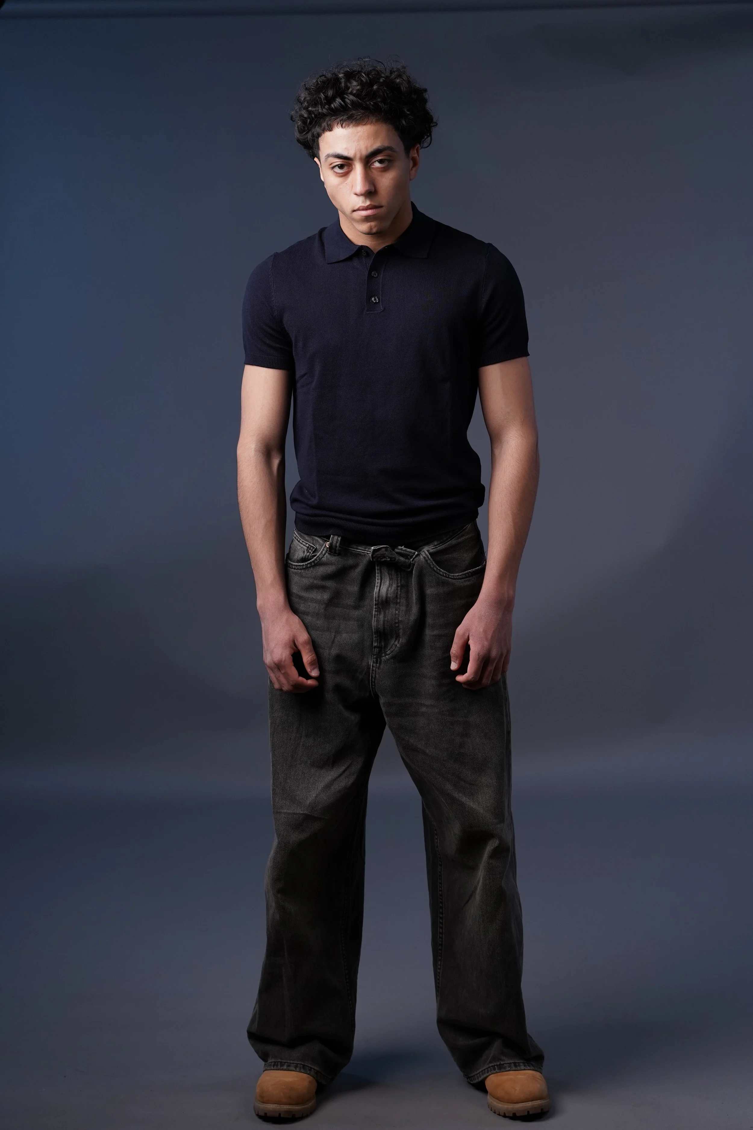 Un giovane uomo con capelli ricci, maglietta nera e jeans neri, che guarda intensamente l'obiettivo, stile studio contro sfondo grigio.