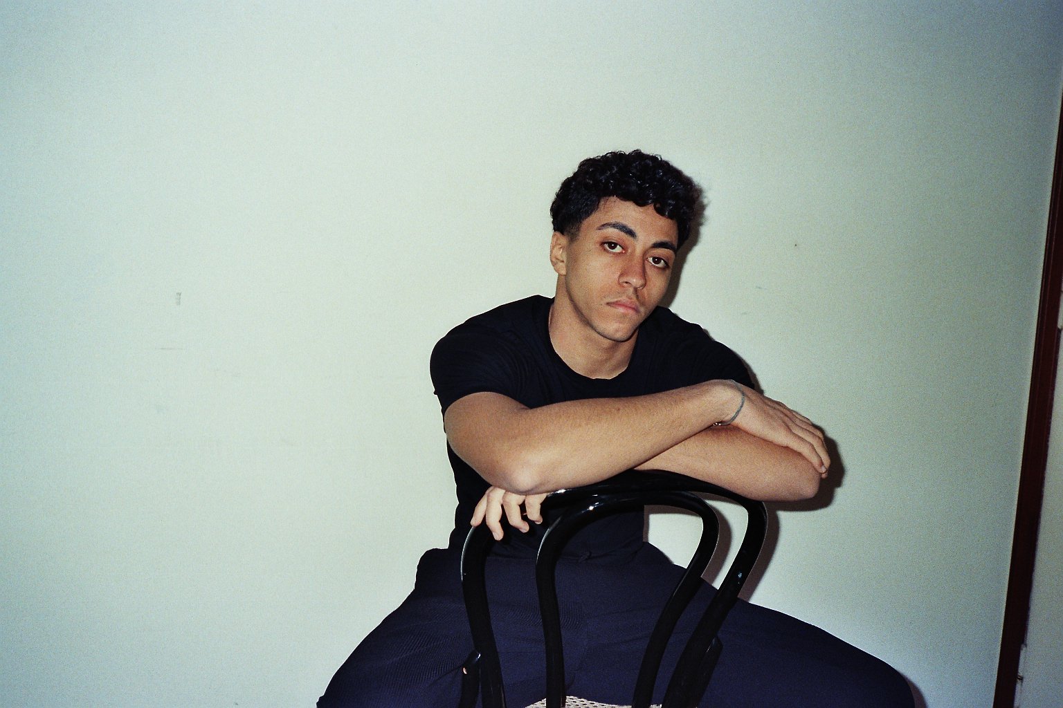 Ragazzo seduto con le braccia appoggiate su una sedia, posa di fronte a una parete chiara.