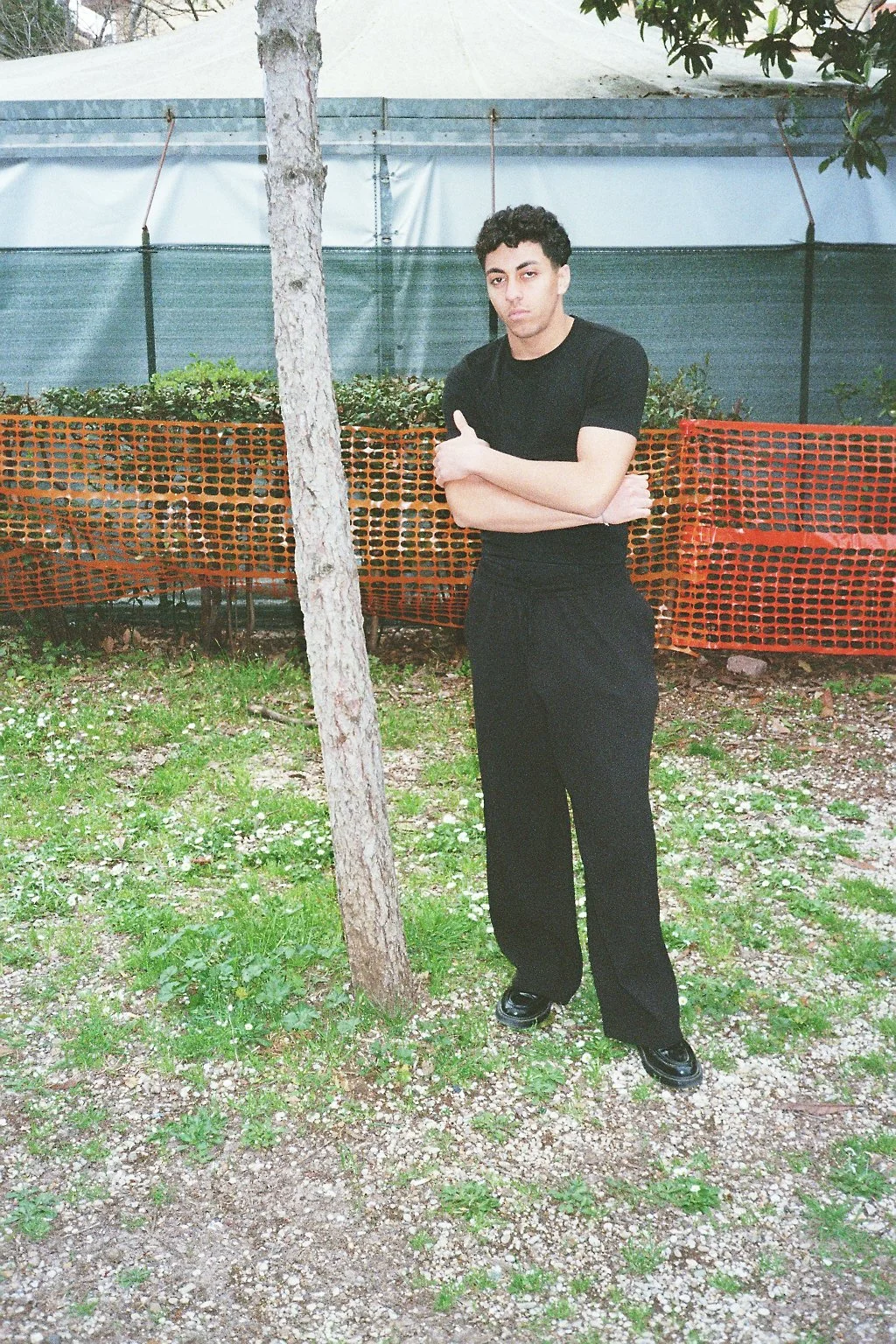 Un giovane uomo vestito di nero, con le braccia incrociate, si trova in un'area esterna con erba e piccoli fiori, dietro una recinzione di rete arancione.