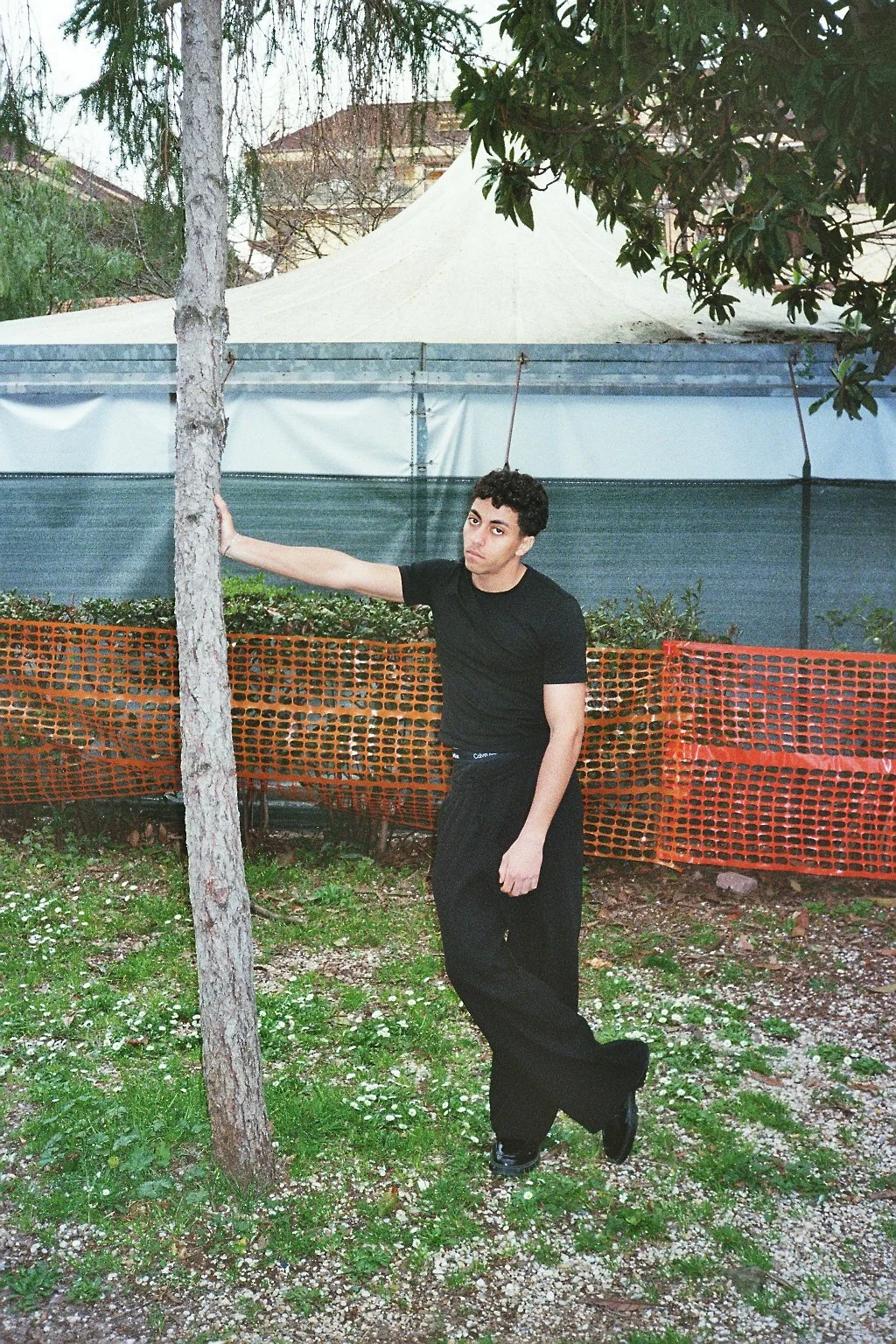 Giovane uomo con capelli ricci, vestito tutto nero, si appoggia a un albero in un'area all'aperto recintata con rete arancione. Sullo sfondo ci sono alcuni alberi e un tendone bianco.