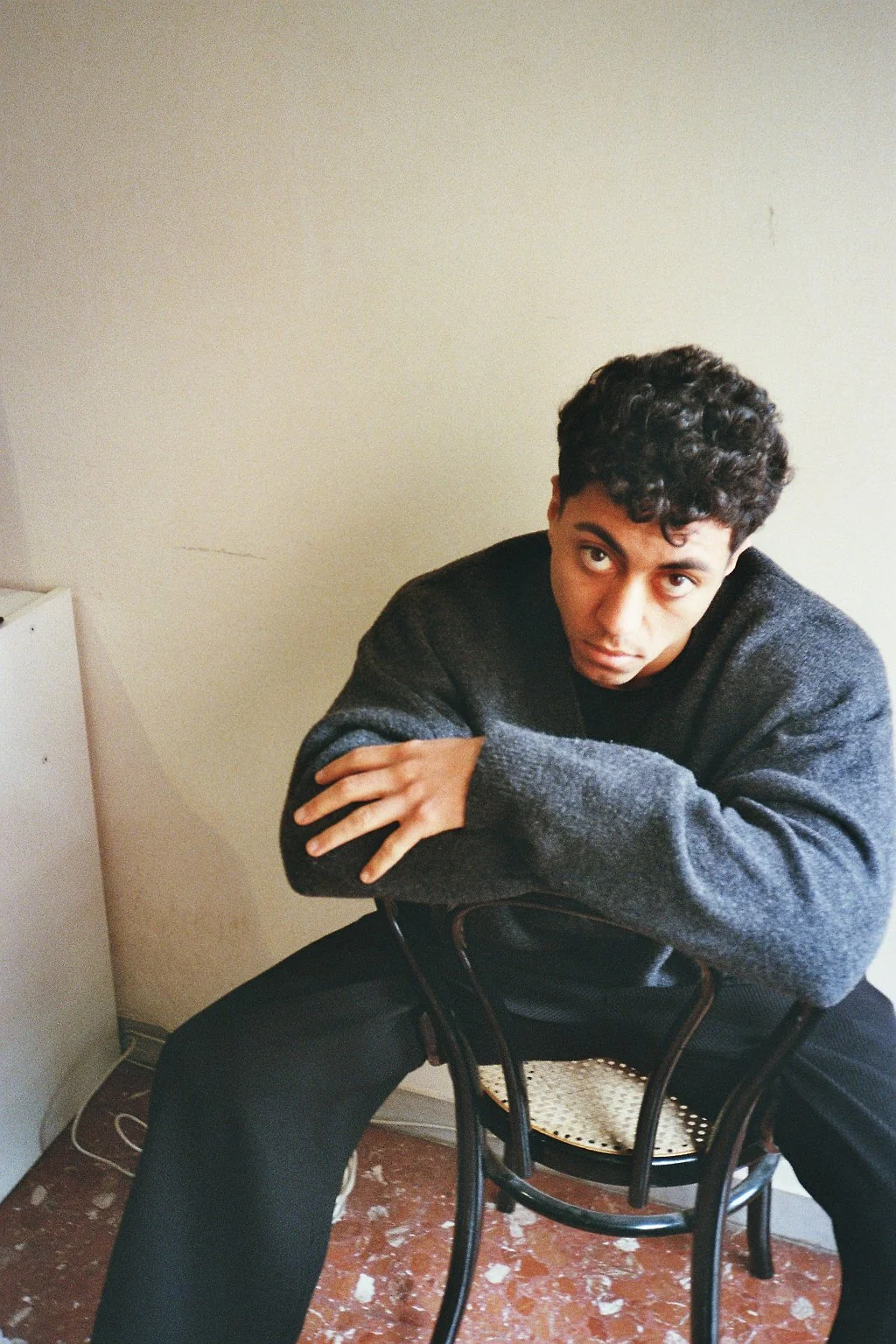 Ragazzo con capelli riccioli seduto su una sedia, indossa un maglione grigio e pantaloni neri, con un'espressione seria, in una stanza con pareti beige e pavimento rosso.