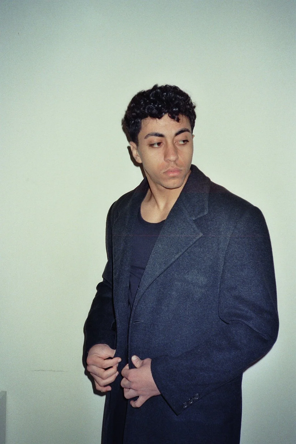 Un giovane uomo con capelli ricciato scuri, indossa un cappotto nero e una maglietta nera, si trova davanti a uno sfondo bianco e guarda leggermente a sinistra.