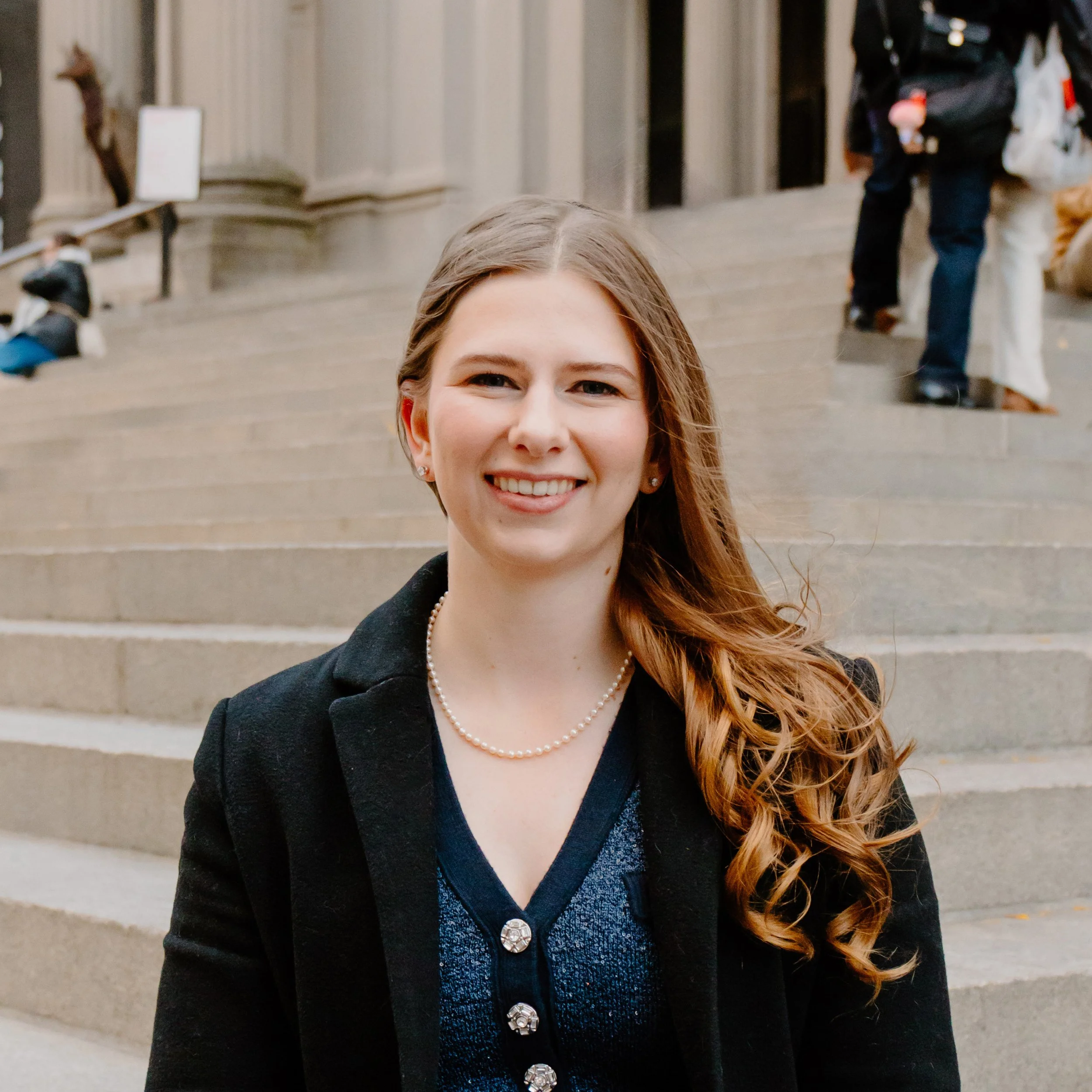 A woman with long, wavy, honey blonde hair smiling in front of a set of stairs outside a building, wearing a black blazer, a dark blue top with decorative buttons, pearl necklace, and small earrings.