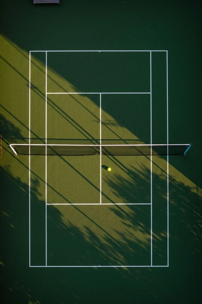 Aerial view of a tennis court with white boundary lines, a net in the middle, a yellow tennis ball near the net, and shadows from surrounding trees.