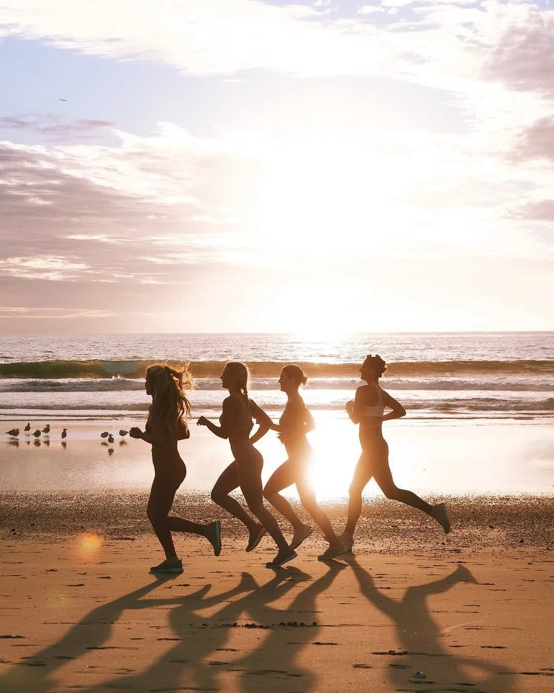 Four women running on the beach during sunset, with shadows cast on the sand, and seagulls near the water.